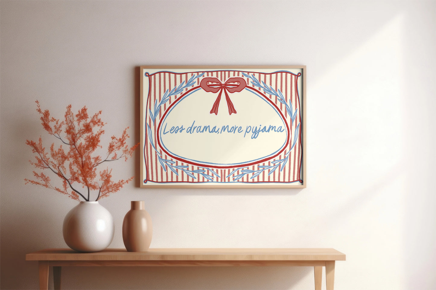landscape orientation print with a beige background, pink stripes and a pink bow with red outline in the top middle. blue outlines of leave form an oval in the center and the quote reads in blue hand writing "less drama more pyjama"