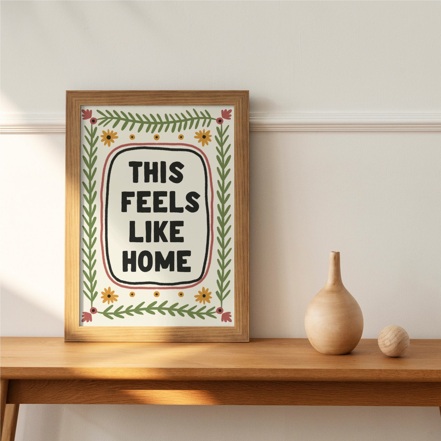 a print shown for display purposes in an oak frame on a wooden table. The print has a quote in the center which reads " this feels like home" in bold black capital letters which are hand drawn. the quote framed by a black inky line and a red one. following the lines of the rectangular, portait image, are folk art style green leafy vines, and 4 yellow flowers accross the top and bottom. in each corner is a red flower. the colour tones are earthy.