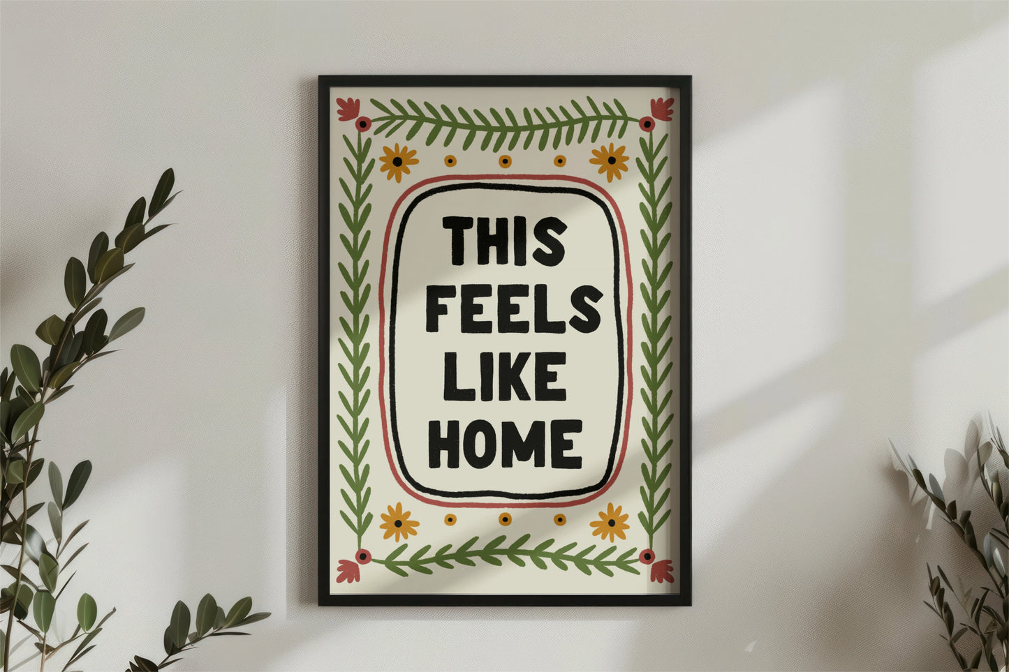 a print shown for display purposes in a black frame on a wall. The print has a quote in the center which reads " this feels like home" in bold black capital letters which are hand drawn. the quote framed by a black inky line and a red one. following the lines of the rectangular, portait image, are folk art style green leafy vines, and 4 yellow flowers accross the top and bottom. in each corner is a red flower. the colour tones are earthy.