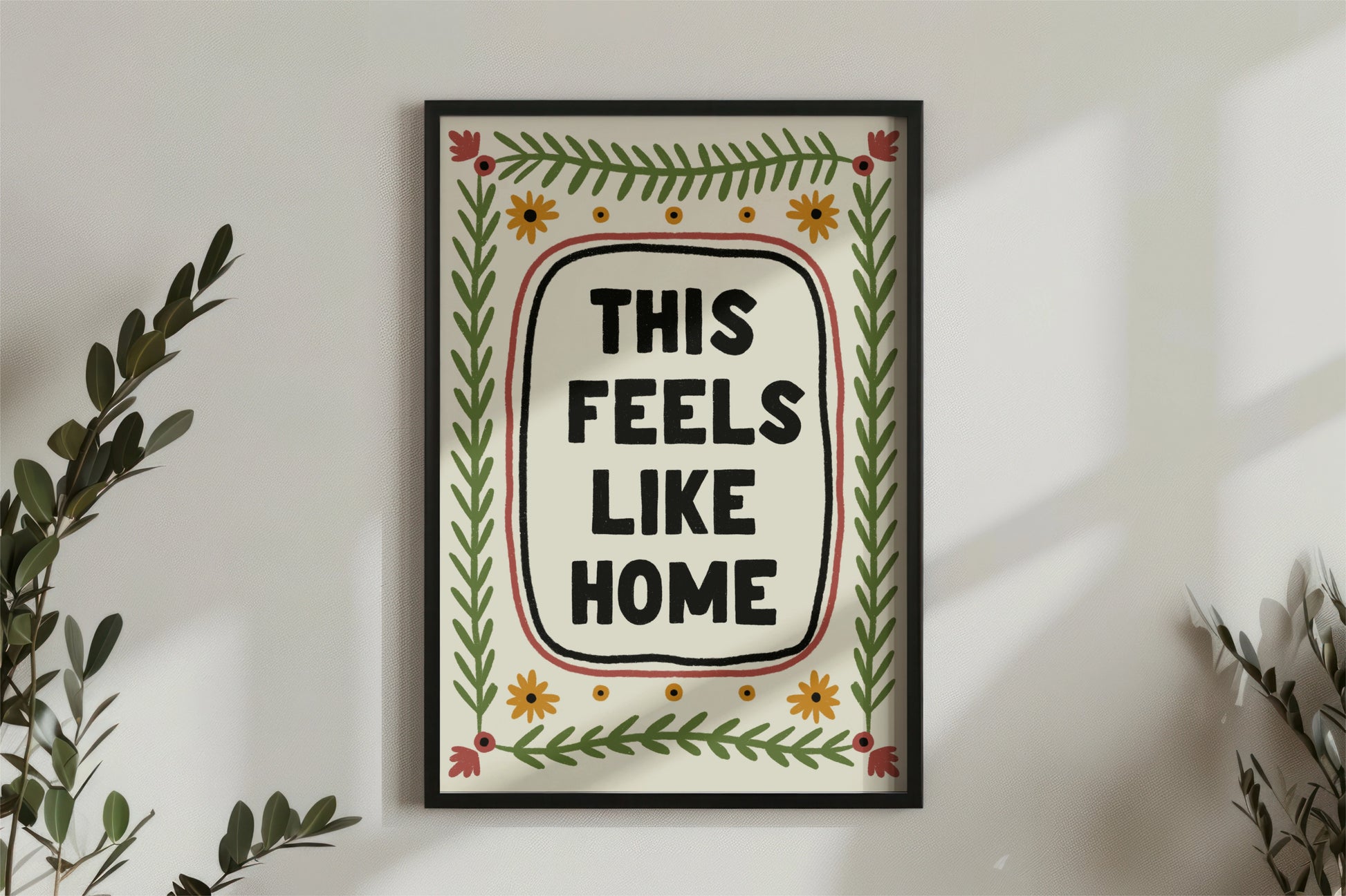 a print shown for display purposes in a black frame on a wall. The print has a quote in the center which reads " this feels like home" in bold black capital letters which are hand drawn. the quote framed by a black inky line and a red one. following the lines of the rectangular, portait image, are folk art style green leafy vines, and 4 yellow flowers accross the top and bottom. in each corner is a red flower. the colour tones are earthy.