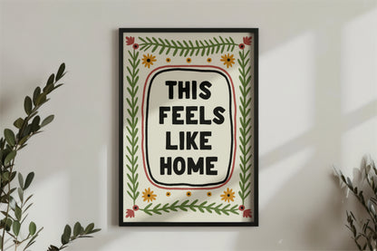 a print shown for display purposes in a black frame on a wall. The print has a quote in the center which reads " this feels like home" in bold black capital letters which are hand drawn. the quote framed by a black inky line and a red one. following the lines of the rectangular, portait image, are folk art style green leafy vines, and 4 yellow flowers accross the top and bottom. in each corner is a red flower. the colour tones are earthy.
