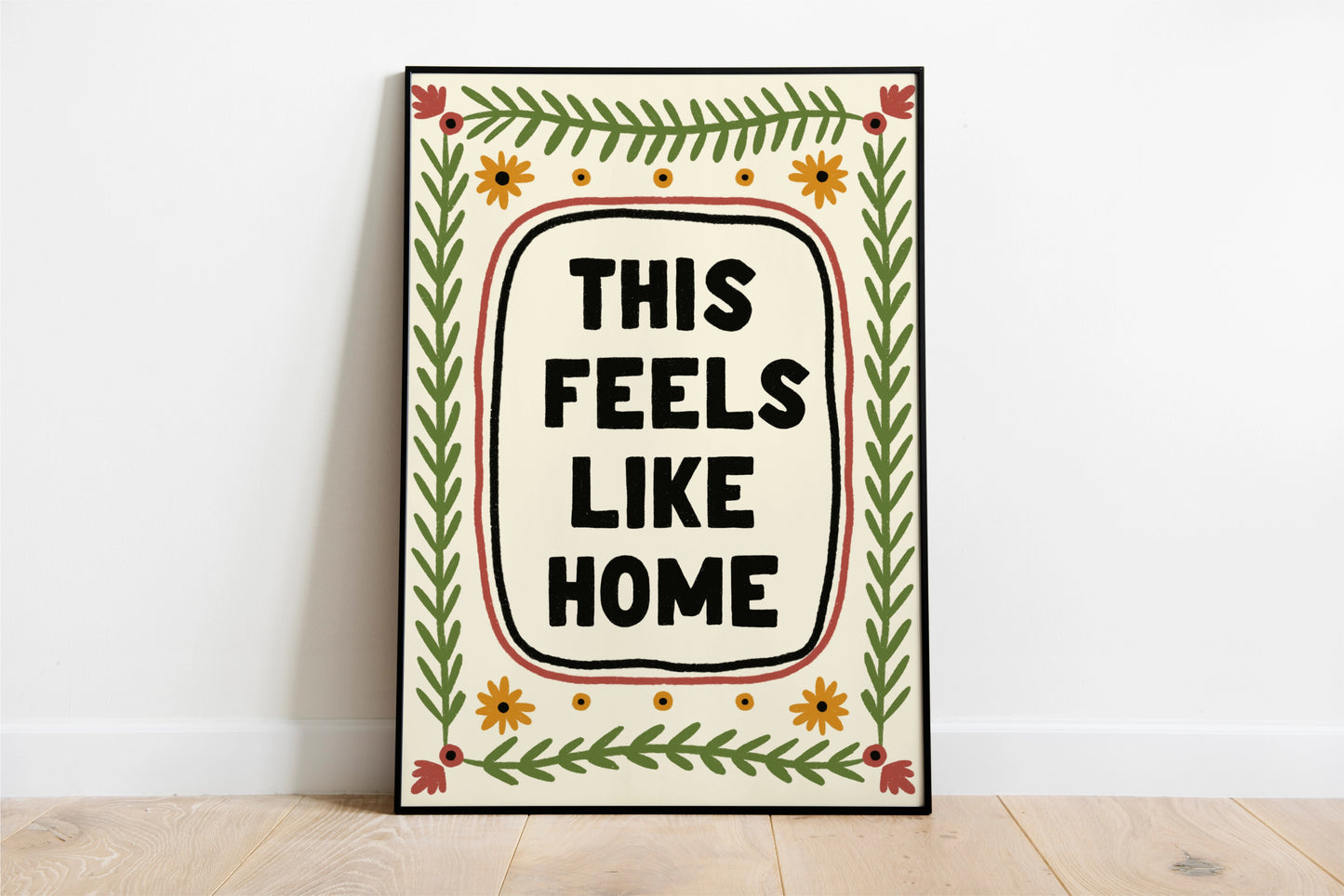 a print shown for display purposes in a thin black frame. The print has a quote in the center which reads " this feels like home" in bold black capital letters which are hand drawn. the quote framed by a black inky line and a red one. following the lines of the rectangular, portait image, are folk art style green leafy vines, and 4 yellow flowers accross the top and bottom. in each corner is a red flower. the colour tones are earthy.