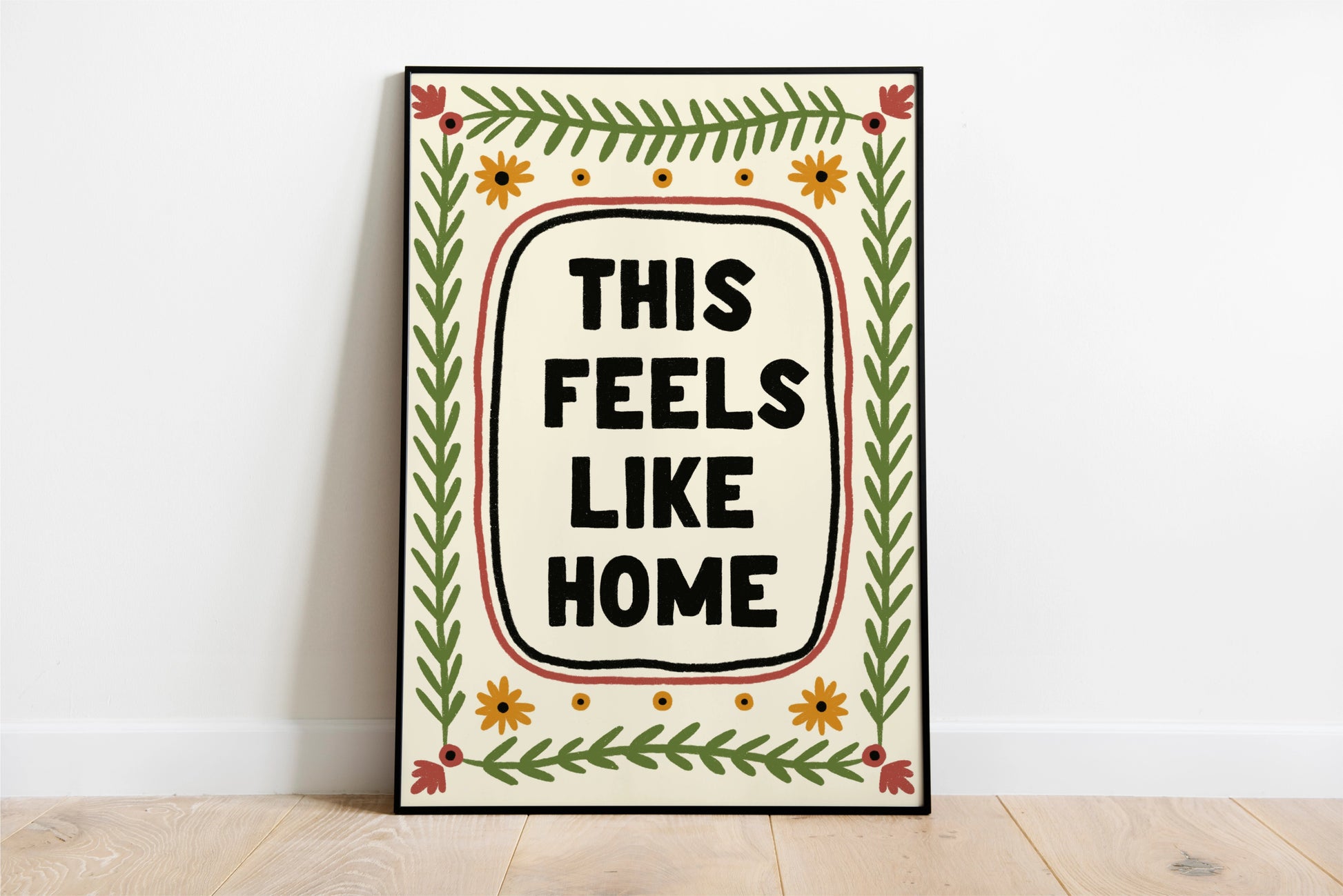 a print shown for display purposes in a thin black frame. The print has a quote in the center which reads " this feels like home" in bold black capital letters which are hand drawn. the quote framed by a black inky line and a red one. following the lines of the rectangular, portait image, are folk art style green leafy vines, and 4 yellow flowers accross the top and bottom. in each corner is a red flower. the colour tones are earthy.